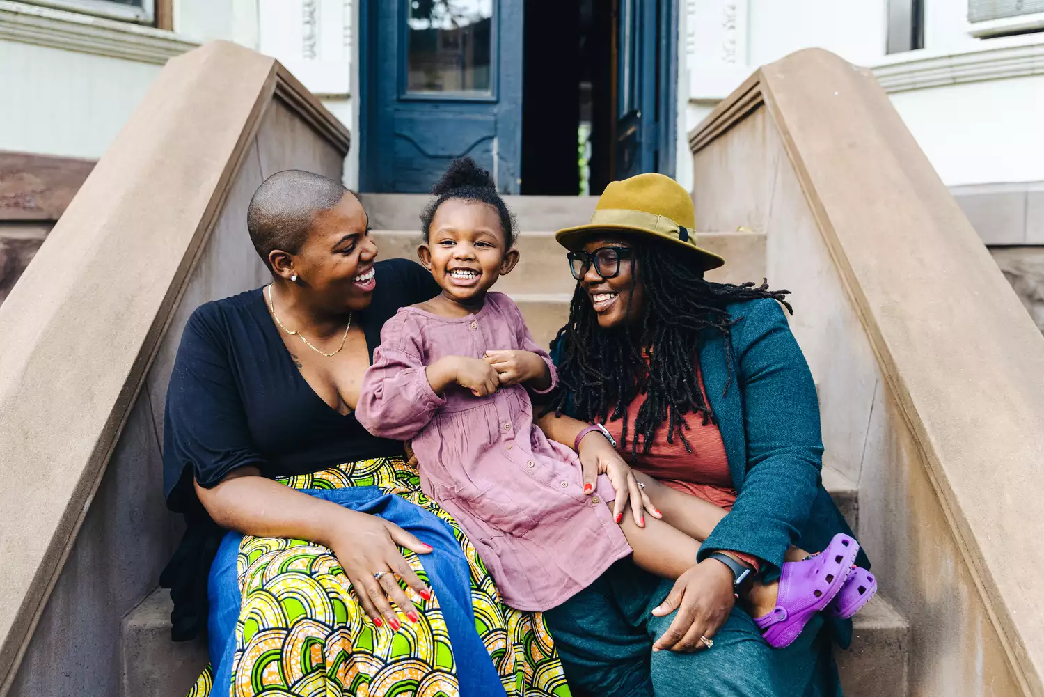 A Black lesbian couple sits on a stoop with their daughter 